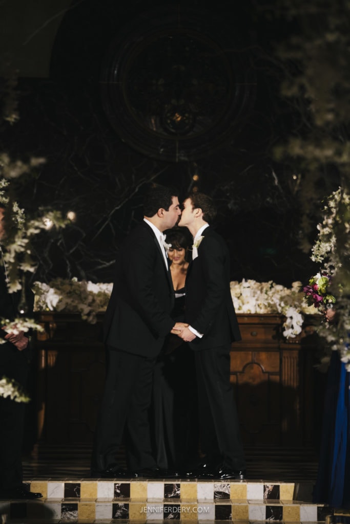 Two grooms are sharing a kiss at their wedding ceremony, both dressed in black tuxedos and holding hands. The background is an elegantly decorated altar with white floral arrangements. An officiant stands behind them. The atmosphere is romantic and celebratory.