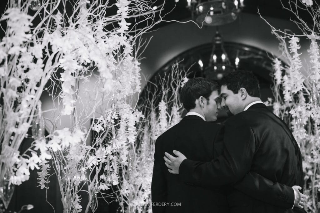 A black-and-white photo of two men in suits sharing an intimate moment, facing each other with arms around one another's backs, surrounded by ornate, white floral decorations. A chandelier hangs above them in a dimly lit environment.