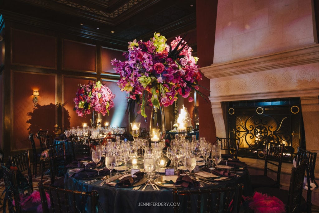A lavish dining setup with elegantly decorated tables featuring large floral centerpieces of pink and purple flowers. The room is dimly lit with warm lighting, showcasing a grand fireplace with intricate metalwork. Glasses, plates, and candles adorn the tables.