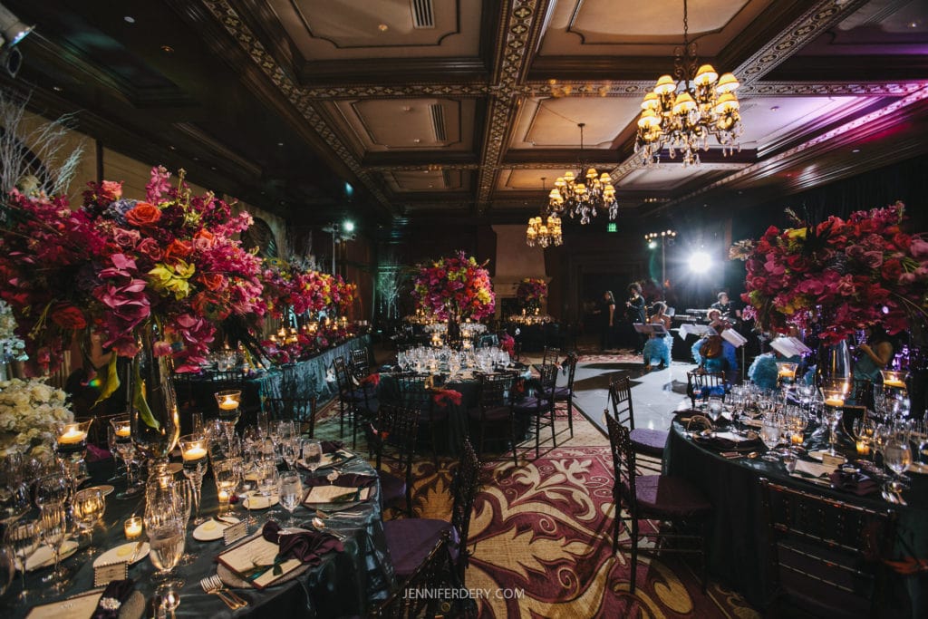 A lavish ballroom is set up for an elegant event. Tables are adorned with towering floral centerpieces in vibrant reds and pinks. Crystal glassware and candles add a touch of sophistication. Chandeliers hang from the ornate ceiling, casting a warm glow over the scene.