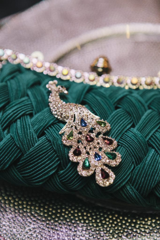 A green textured handbag adorned with a detailed, jewel-encrusted peacock brooch. The peacock's wings are embedded with multicolored gemstones, adding a touch of elegance to the accessory. The background features a shimmering surface.
