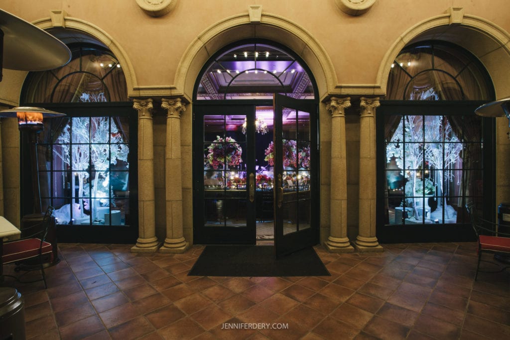 A grand entrance with ornate columns and three large arched glass doors lit by a purple light. Inside, a room is decorated with lush, colorful floral arrangements and ambient lighting. The interior conveys an elegant and festive atmosphere.