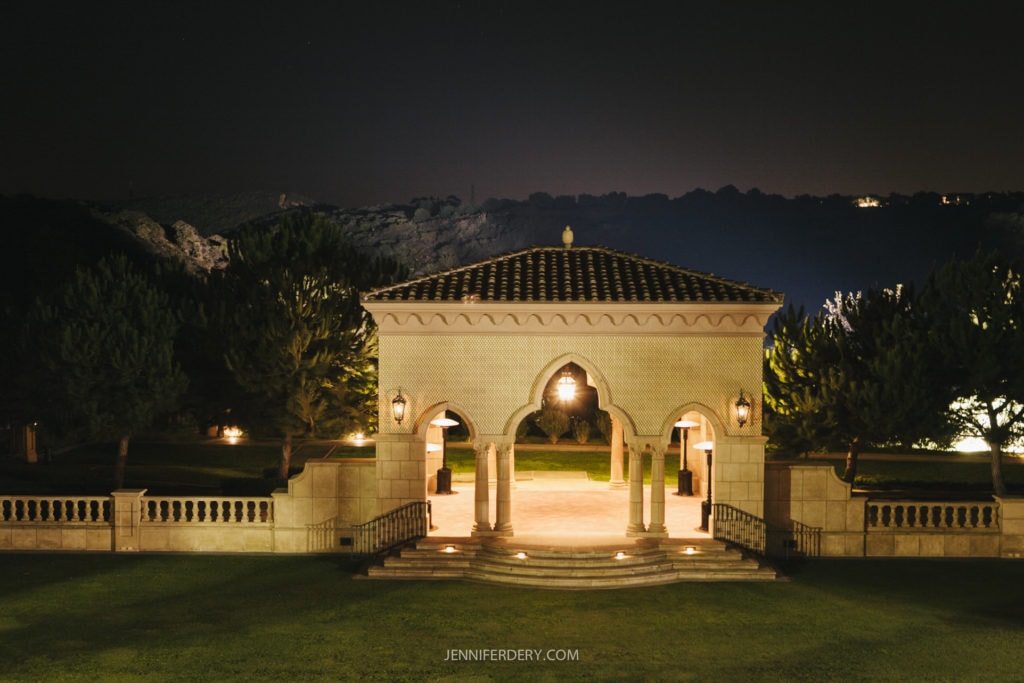 A night view of a beautifully lit, small pavilion at the grand del mar with an arched entrance, surrounded by manicured lawns and trees. The structure has a tiled roof, glowing warmly, with a hilly landscape visible in the background under a dark sky.