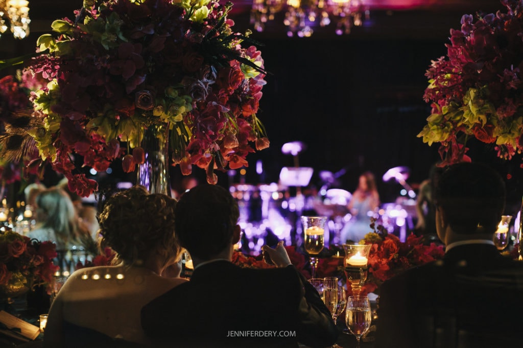 A dimly lit event features elegantly dressed guests seated at a table adorned with large floral arrangements and warm candles. The background is a blurred stage with musical instruments, suggesting a live performance or concert atmosphere.