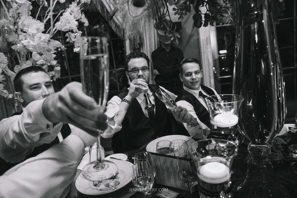 A group of men in formal attire are seated around a table, raising their glasses in a toast. The table is adorned with candles, glasses, and floral arrangements. The mood appears celebratory and jovial. The image is in black and white.