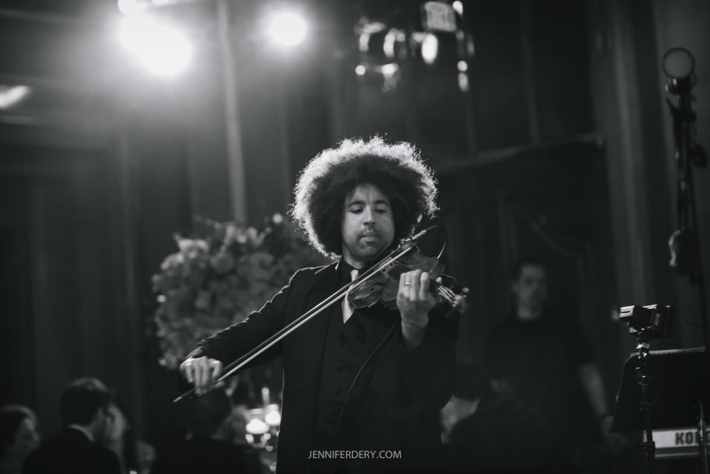A musician with curly hair, dressed in a suit, passionately plays the violin in a dimly lit setting. The background is blurred, with a few people and decorative elements visible. The scene appears to be a formal event or performance.