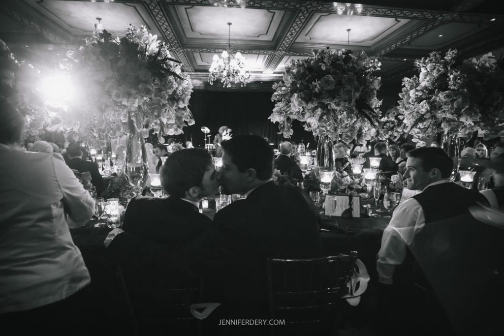 A black-and-white photo captures two men sharing a kiss at a lavishly decorated event with large floral arrangements and elegant chandeliers. Other guests are mingling around the table, creating a warm and celebratory atmosphere.