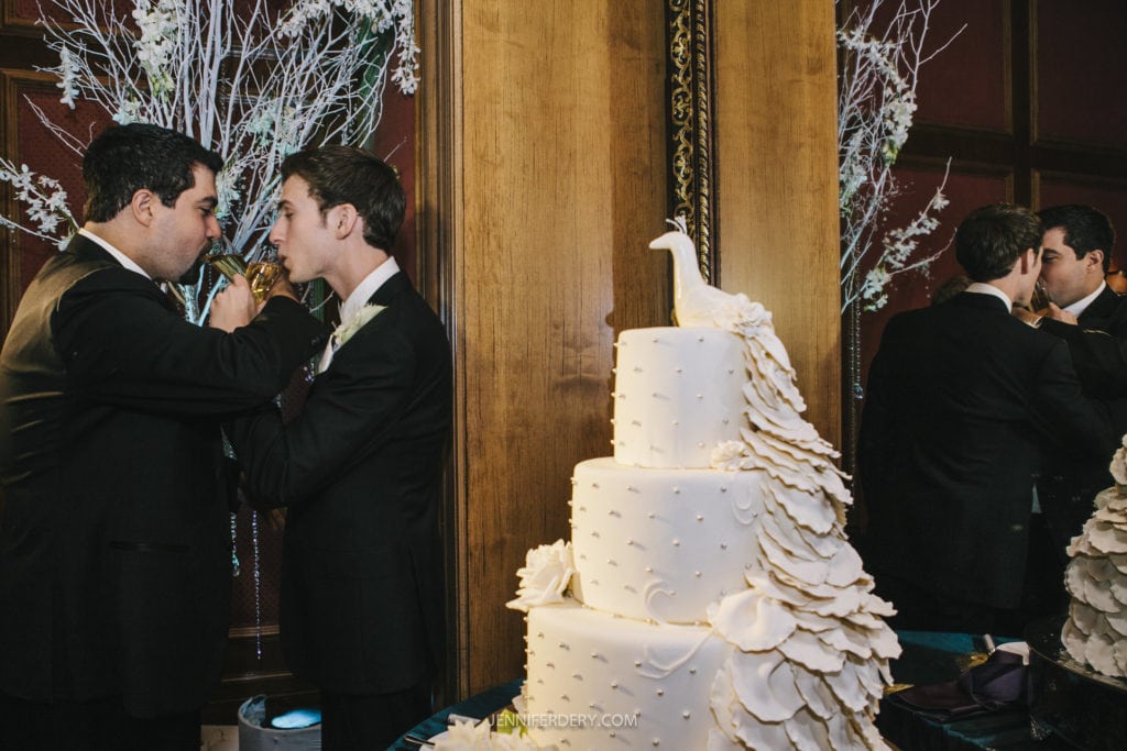 Two men in formal black suits are celebrating a wedding. They are drinking from champagne flutes, arms interlinked. In the foreground, a tiered wedding cake decorated with a white peacock detail is visible. White floral arrangements are in the background.