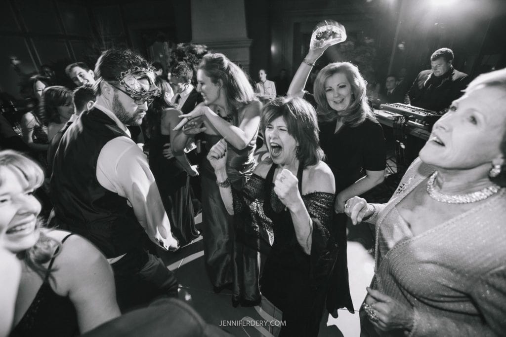 A black and white photo shows a lively crowd of people dancing at an indoor event. Many are smiling and cheering. Some wear masks, adding a festive touch to the gathering. The background features a DJ booth and atmospheric lighting enhancing the celebratory ambiance.