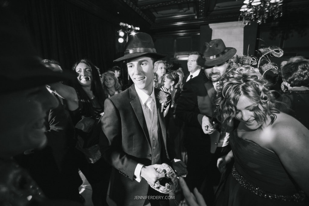 A black-and-white photo shows people in formal attire dancing at an event. A man in the center wears a suit and hat, smiling towards the camera. A woman in a dress dances nearby, along with other attendees in the background. The atmosphere is festive and lively.