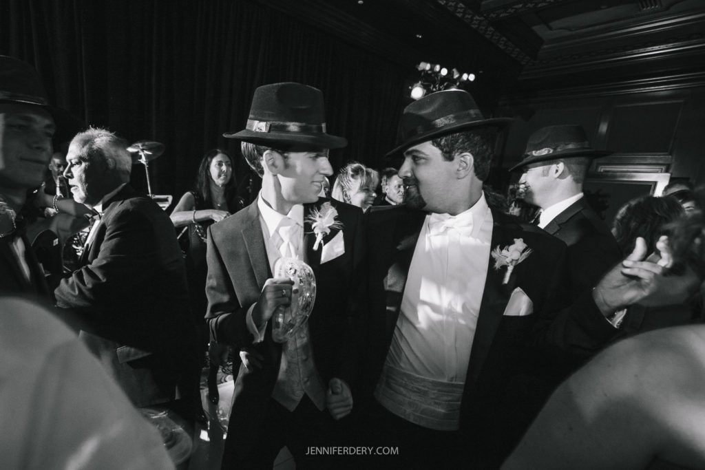 A black-and-white photo of two men in suits, wearing fedora hats, facing each other and smiling. One man is holding a tambourine. They are surrounded by other people wearing formal attire at what appears to be a lively event or celebration.