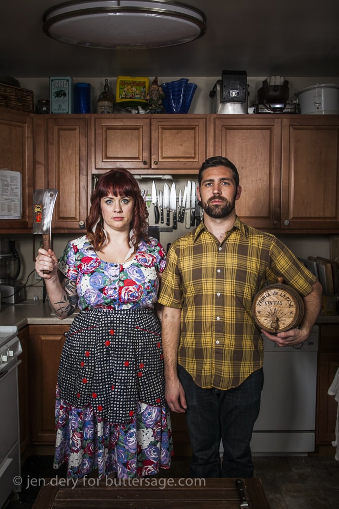 A woman in a colorful floral dress and apron sternly holds a cleaver while standing next to a man in a yellow plaid shirt holding a wooden barrel. They are in a kitchen with wooden cabinets. The setting and their expressions give a humorous, posed appearance.