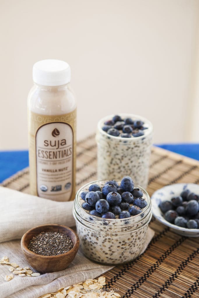 A bottle of Suja Essentials Vanilla Nut beverage stands behind two jars of chia pudding topped with blueberries. A small wooden bowl filled with chia seeds and additional blueberries in a dish are placed in the foreground on a woven mat.