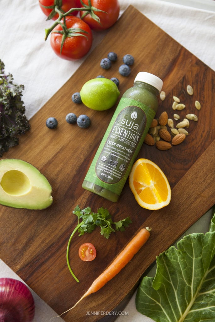 A wooden cutting board featuring a bottle of Suja Essentials green juice surrounded by fresh fruits and vegetables, including an avocado half, tomatoes, blueberries, orange slice, carrot, lime, almonds, kale, and a sprig of parsley.