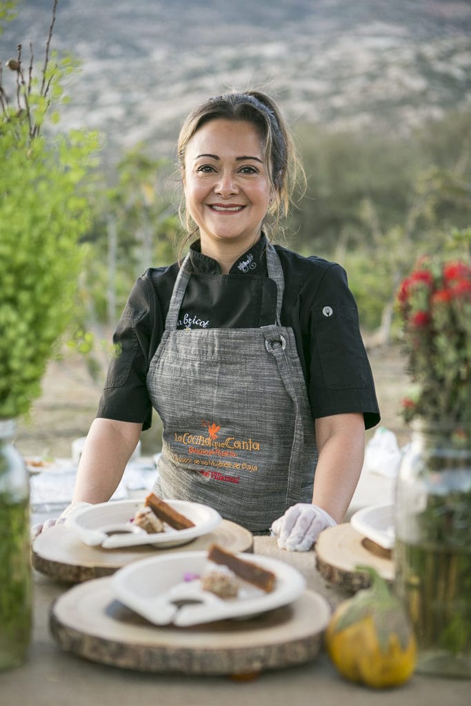 A woman wearing a black shirt and a gray apron stands at an outdoor table with desserts elegantly plated on wooden boards. She smiles at the camera, with greenery and a mountainous landscape in the background.
