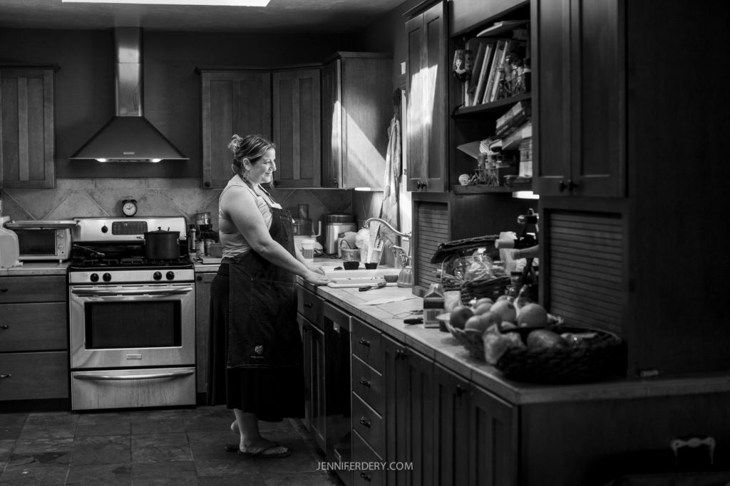 Chef Katie Grebow stands in her kitchen, wearing an apron, and working at the sink. The kitchen is well-equipped with cabinets, a stove, and various items on the countertops and shelves. The scene is lit softly, creating a cozy atmosphere. The image is in black and white.