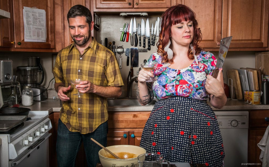 A man and a woman are standing in a kitchen. The man, wearing a yellow plaid shirt, holds a drink and is smiling. The woman, wearing a floral top and polka dot skirt, holds up a large knife while looking at it intently. Various kitchen utensils hang in the background.