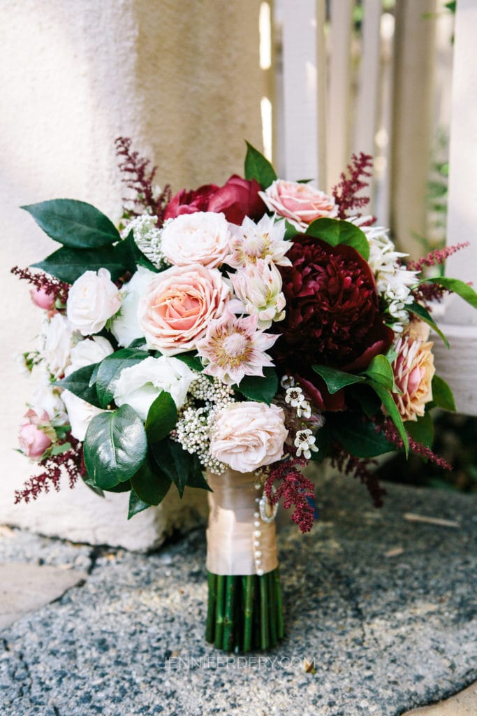 A charming bridal bouquet resting against a white fence, featuring an array of pink, peach, and deep red flowers, including roses and peonies. The bouquet is accented with green leaves and small white flowers, with stems wrapped in a beige ribbon adorned with pearls.