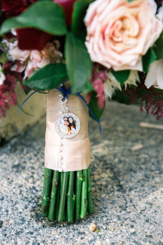 Close-up of a wedding bouquet with a mix of blush, burgundy, and white flowers. The stems are wrapped in cream-colored satin ribbon, adorned with pearl-like buttons and a circular locket pendant with a photo of a couple attached with a blue ribbon.