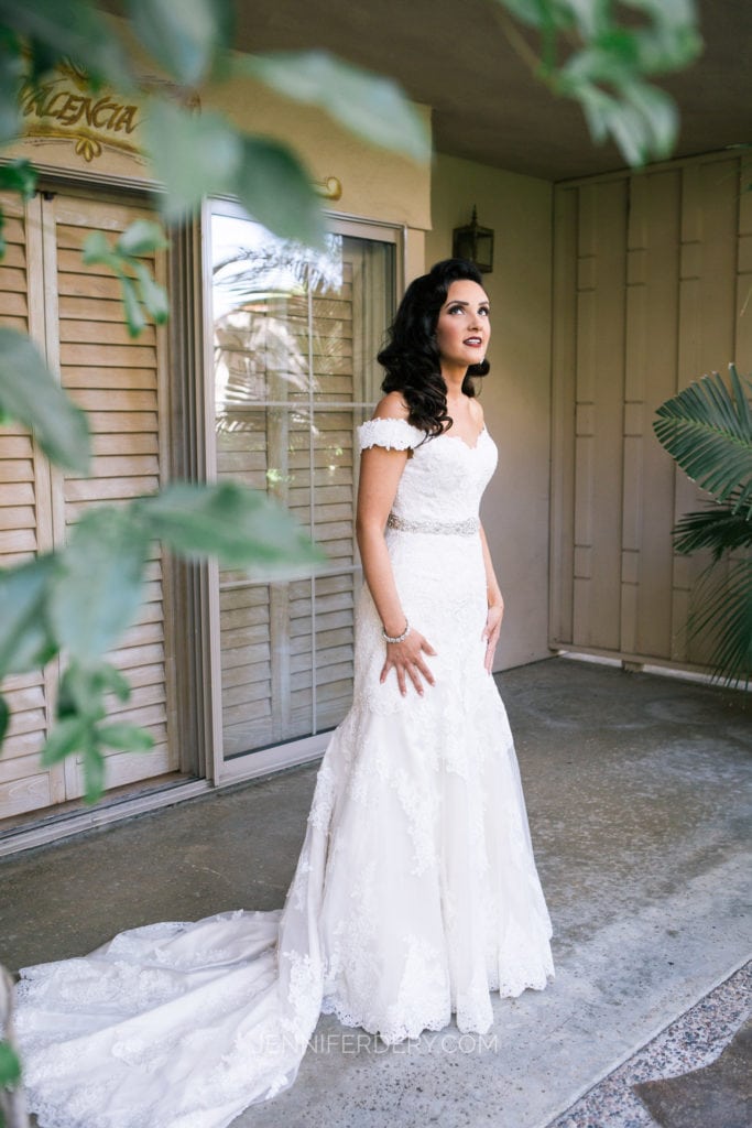 A bride stands in front of a house, dressed in a white off-the-shoulder wedding gown with intricate lace details and a long train. She looks off to the side with a thoughtful expression, surrounded by green foliage and next to a glass patio door and shutters.