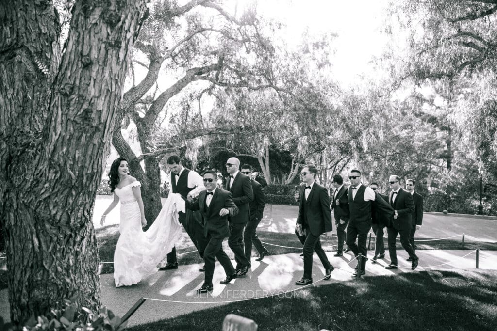 A black-and-white photo showcasing a bride in a long gown being escorted by groomsmen and a group of men in suits. They are walking along a path surrounded by trees and greenery, with sunlight filtering through the branches.
