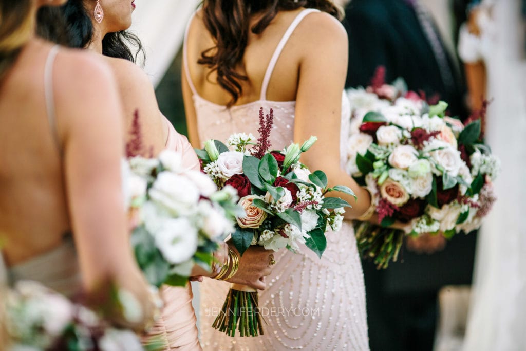 Three women in light-colored dresses hold bouquets of flowers with white, red, and green hues. The focus is on the floral arrangements and their elegant attire, while the background is slightly blurred.