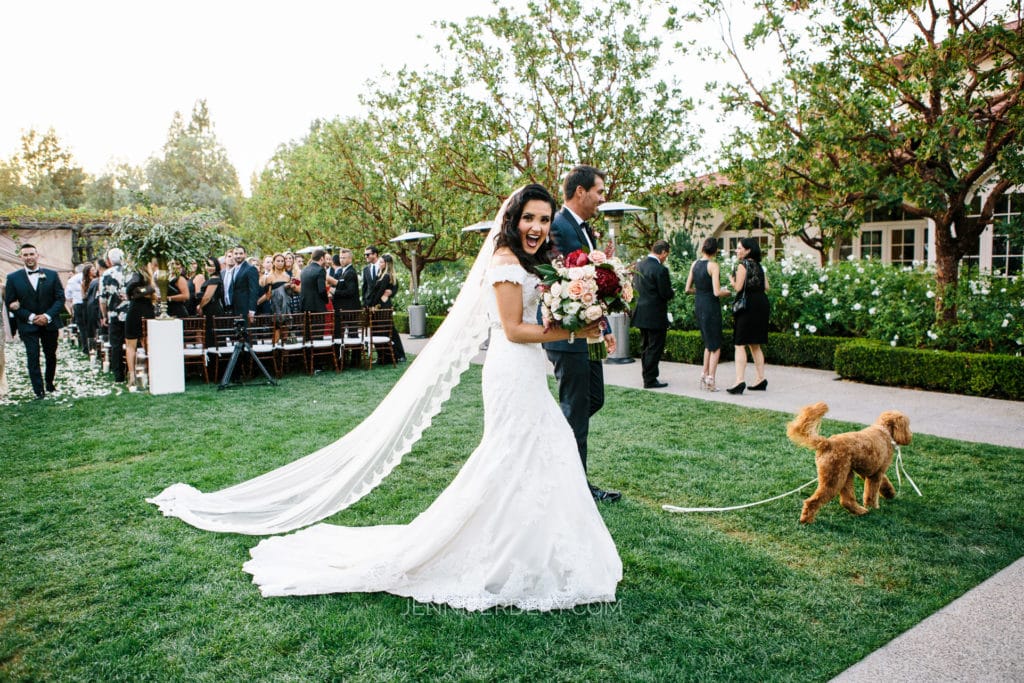 A bride in a white dress and long veil walks joyfully through a garden aisle at Rancho Bernardo Inn holding a bouquet as the groom follows, grasping a leash of a brown poodle. Guests in formal attire are seen in the background, seated and standing, amidst greenery and floral arrangements.
