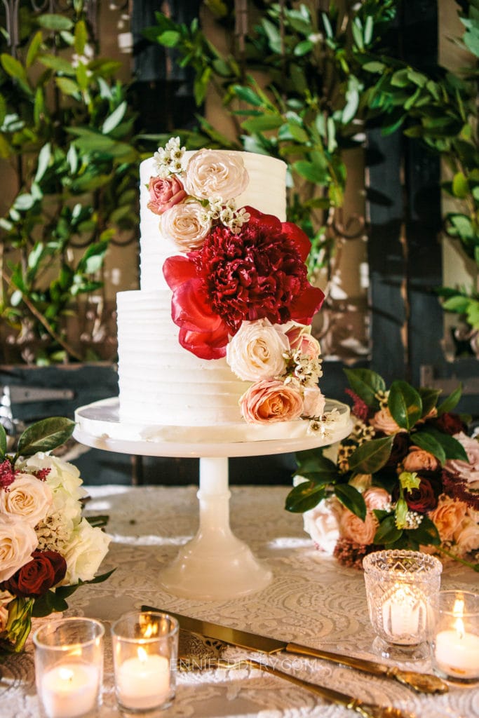 A tiered white wedding cake adorned with red and blush flowers, placed on a white cake stand. The table is decorated with candles in glass holders and floral arrangements, set against a backdrop of greenery.