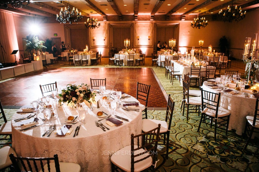 A spacious wedding reception hall with round tables covered in white linens, elegantly set with glassware, napkins, and floral centerpieces. The room is softly lit, featuring chandeliers and ambient lighting. A large, open dance floor is centered in the space—perfect for communicating your vision to your wedding photographer.
