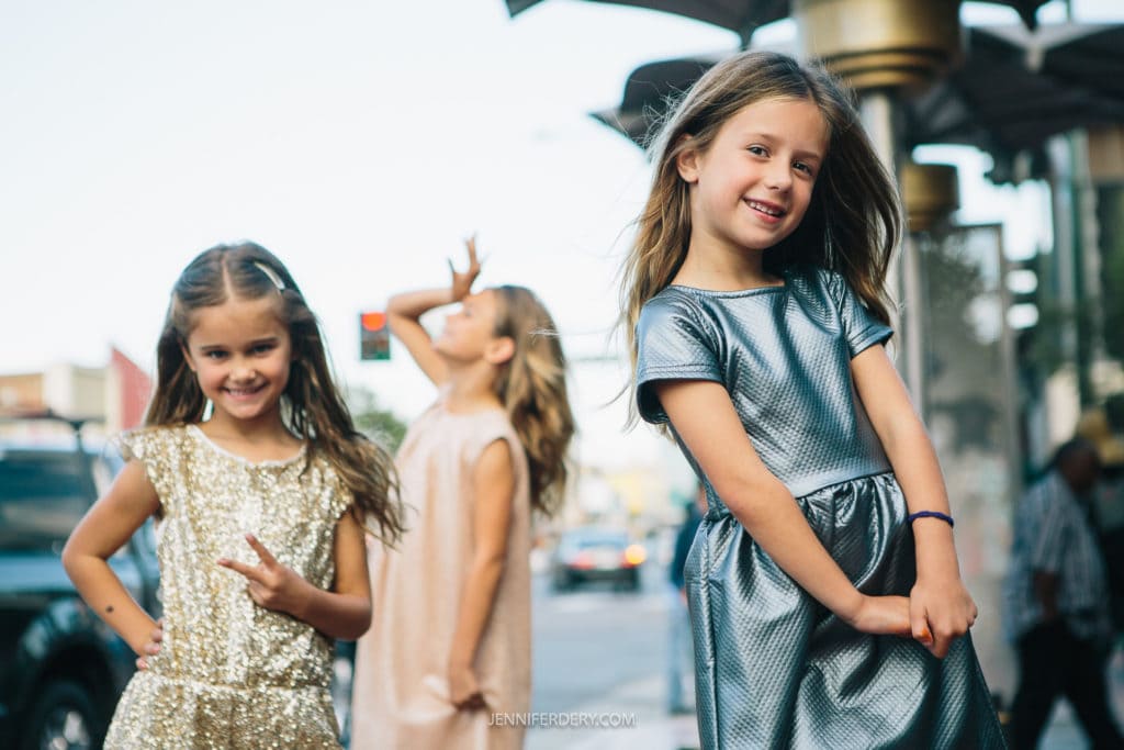 Three young girls are posing outdoors. The girl in front is wearing a metallic silver dress and smiling at the camera, while the girl to the left is in a gold dress, making a peace sign. The third girl, in a blush dress, stands in the background with her hand raised.