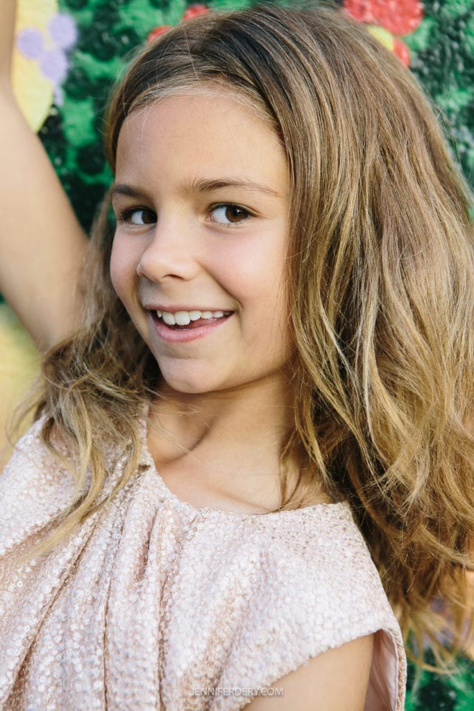 A young girl with long, light brown hair smiles while standing in front of a colorful, floral-patterned wall. She wears a sleeveless, light textured top.