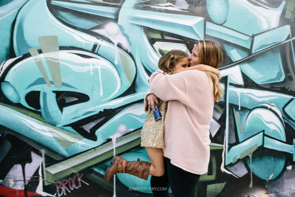 A woman in a light sweater joyfully hugs a young girl in a sparkling dress and brown boots. They are standing in front of a colorful graffiti wall with shades of blue and black.