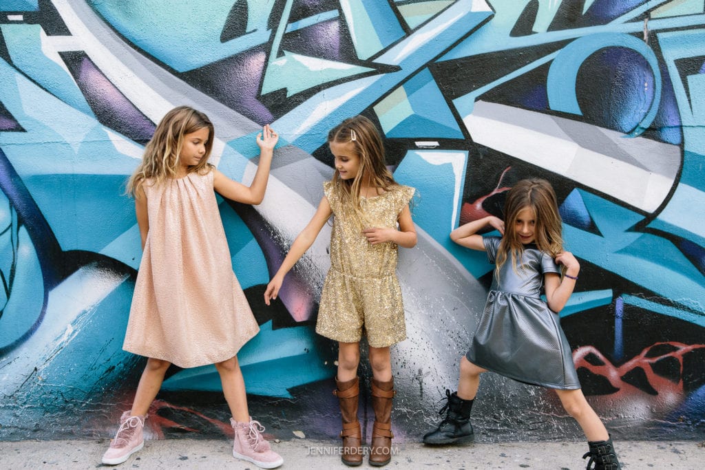 Three young girls strike playful poses in front of a vibrant graffiti wall. They are wearing shimmering dresses in shades of pink, gold, and silver, paired with boots. The art on the wall features abstract shapes in blue and purple tones.