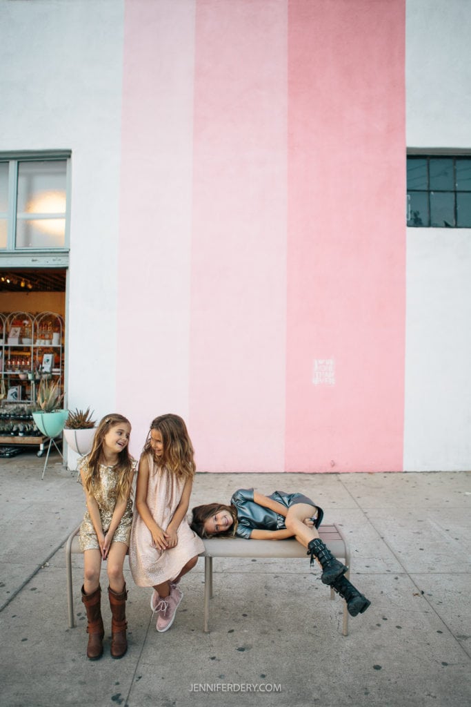 Three girls pose in front of a building with a wide vertical pink stripe. Two of the girls sit on a bench, smiling and chatting, while the third girl reclines on the bench, one leg propped up, looking relaxed. The setting appears to be urban with an open door to a shop.