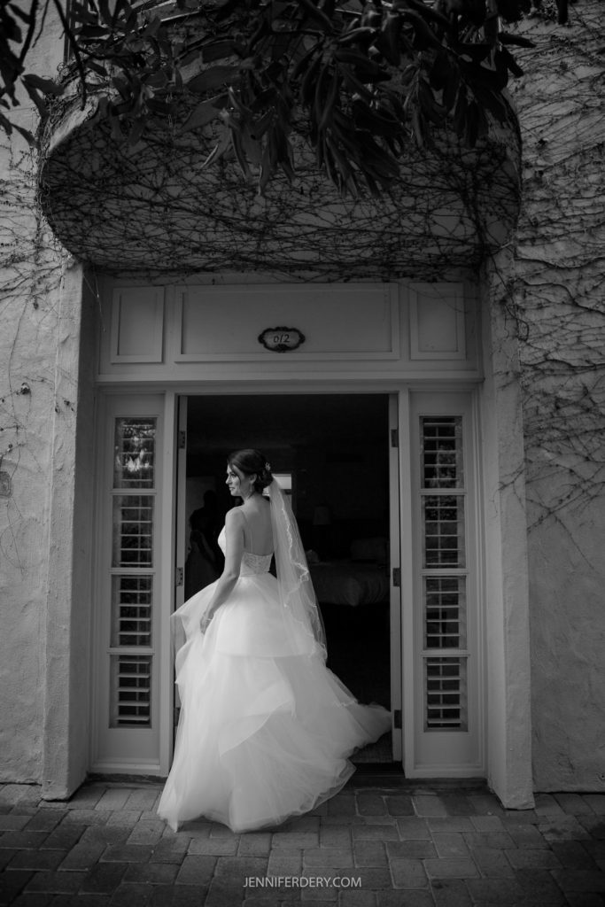 black and white full-length image of a bride standing outside her hotel room checking out the rain on her wedding day