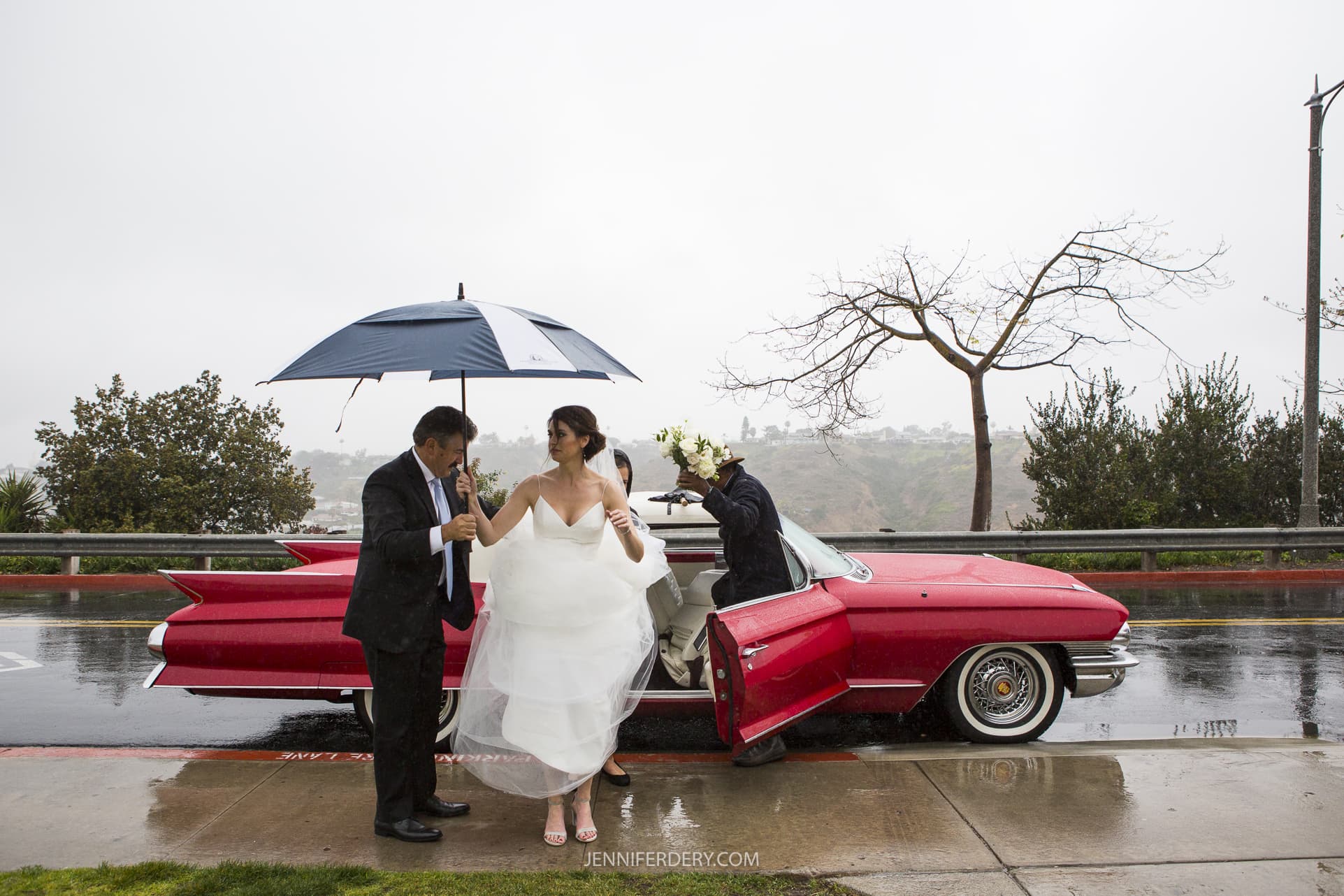 bride exiting a red convertible classic car in the rain. she is wearing a long white wedding dress, the car is parked on a wet road and her dad is helping her with a big golf umbrella