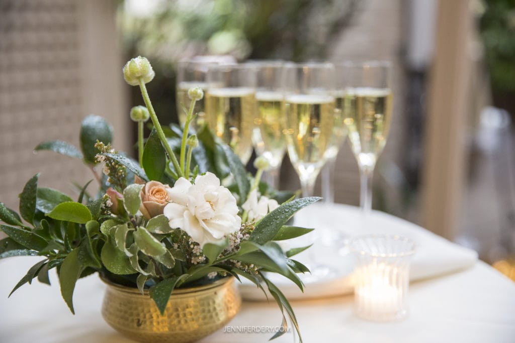 wedding floral decor cocktail table greens and flowers in a gold bowl with champagne flutes and a candle in the background
