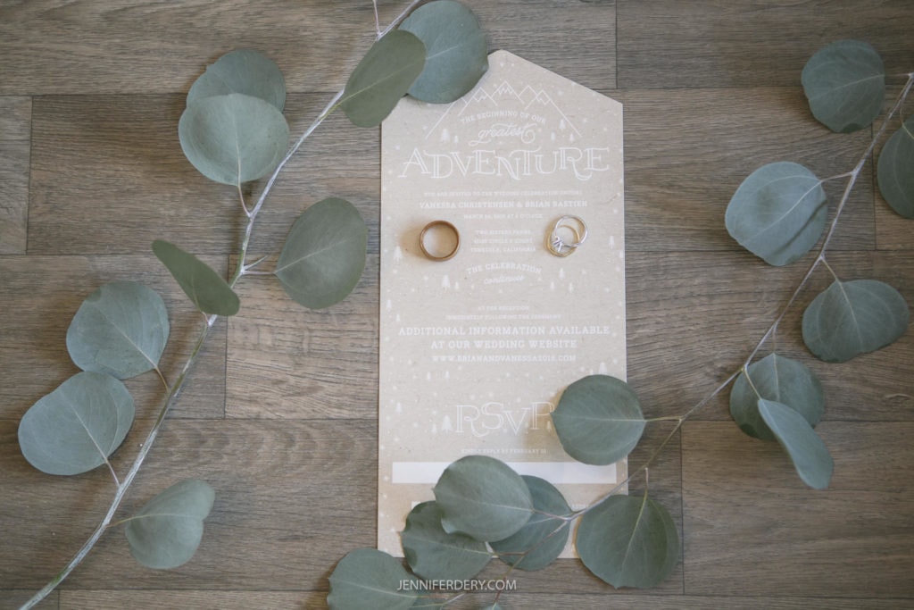 A wedding invitation lies on a wooden surface, adorned with green eucalyptus leaves. Two wedding rings are placed on the invitation, which features a white mountain-themed design and text providing event details. "RSVP" is visible at the bottom, enhancing the rustic wedding charm.
