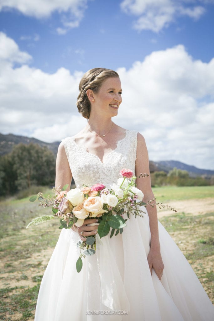 A bride in a white wedding dress stands outside on a sunny day, smiling and holding a bouquet of flowers. The background features a scenic landscape with mountains and a cloudy blue sky, perfectly capturing the charm of a rustic wedding.