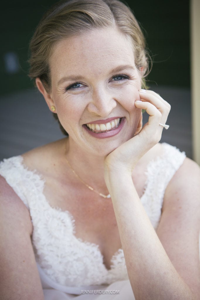 A smiling bride in a white lace wedding dress rests her head on her hand, showcasing her ring. She has light brown hair and is posing outdoors against a dark green blurred background, perfectly embodying the charm of a rustic wedding.