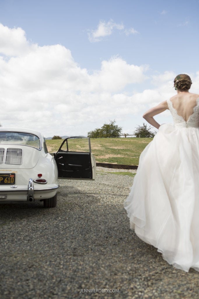 vintage car with door open. a bride in a long white dress is walking towards it. Photo communicates the vision by the Wedding Photographer of a bride getting ready to drive to her wedding