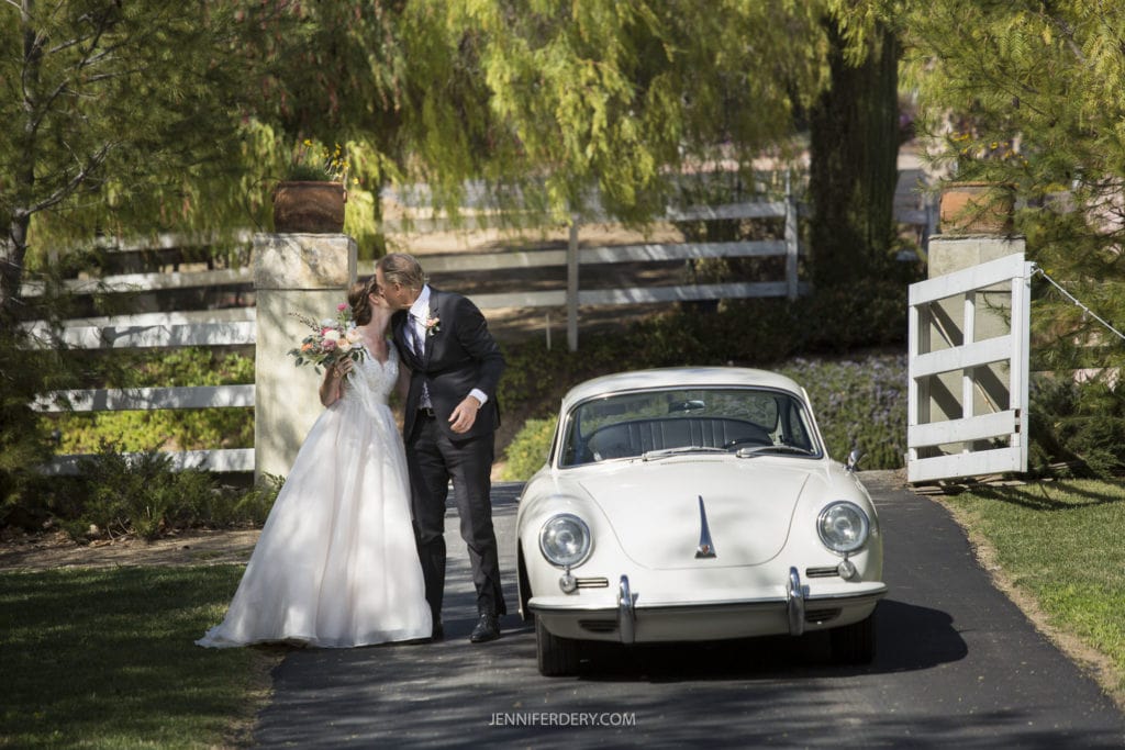 A newlywed couple shares a kiss outside by a white vintage car, surrounded by lush greenery. The bride, in a white gown and holding a bouquet, and the groom, dressed in a black suit, epitomize the charm of their rustic wedding. A white fence and gate are visible in the background.