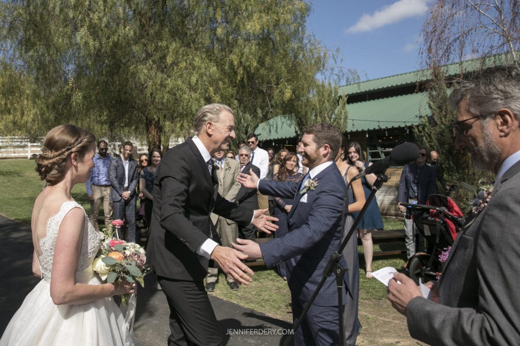 A groom and bride in wedding attire stand outdoors with a group of guests, celebrating a charming rustic wedding. An older man in a black suit reaches out to hug the groom. The background features green trees and a green-roofed building under a clear blue sky.