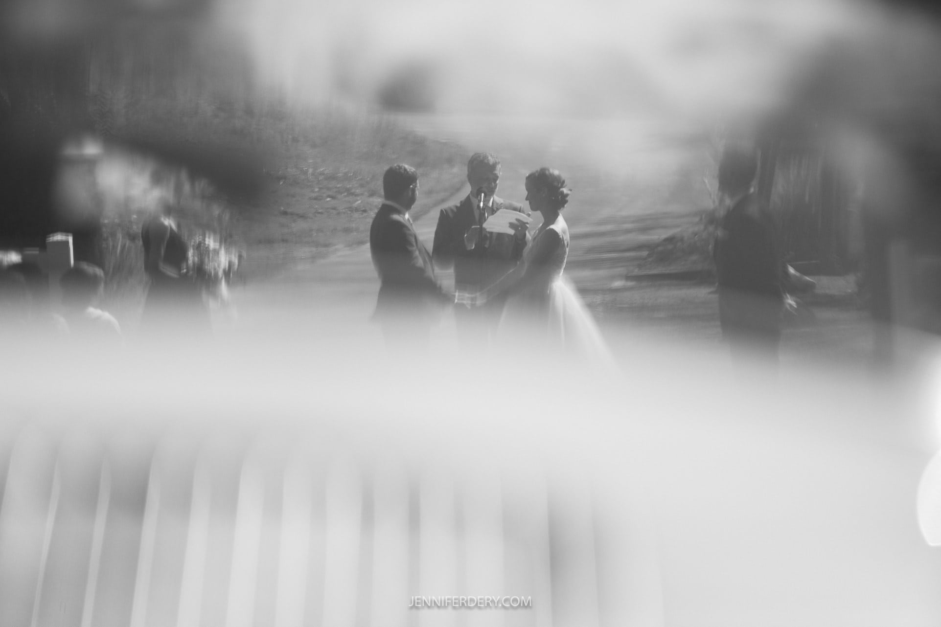 A black and white image shows a bride and groom standing before an officiant outdoors at a rustic wedding. The scene is blurred and softly focused, creating a dreamy effect. The couple appears to be exchanging vows or listening to the officiant.