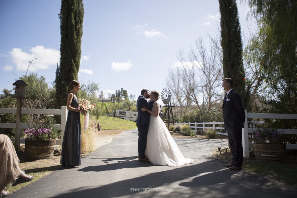 A newlywed couple shares a kiss during their outdoor, rustic wedding ceremony on a sunny day, flanked by two tall trees. The bride wears a white gown while the groom is in a suit. Two attendants stand nearby, one holding a bouquet. A windmill and white fence add charm to the backdrop.