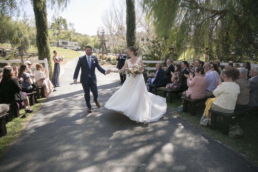 A newly married couple joyfully walks down an outdoor aisle lined with seated guests who are clapping and cheering. The bride wears a white wedding dress and holds a bouquet, while the groom is in a suit. The rustic wedding scene is set in a garden with trees and a windmill in the background.