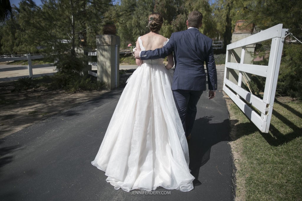A bride in a white gown and a groom in a blue suit walk arm in arm down a paved path surrounded by greenery and a white fence, capturing the charm of their rustic wedding.