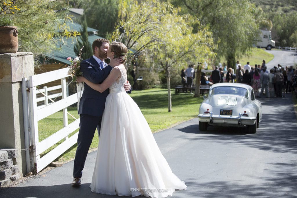 A bride and groom share a kiss beside a white fence on a sunny day, with the groom holding a bouquet. They stand near a vintage car parked on the road, embodying the charm of a rustic wedding, while a group of people gathered in the background suggests an ongoing celebration.