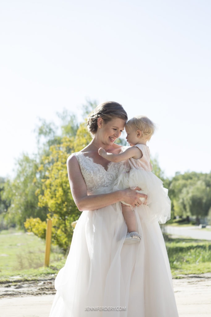 A woman in a white dress is holding a small child in a matching white dress. They are outdoors with trees and greenery in the background, reminiscent of a rustic wedding. The woman is looking affectionately at the child, and both are smiling. The sun is shining brightly, creating a serene atmosphere.