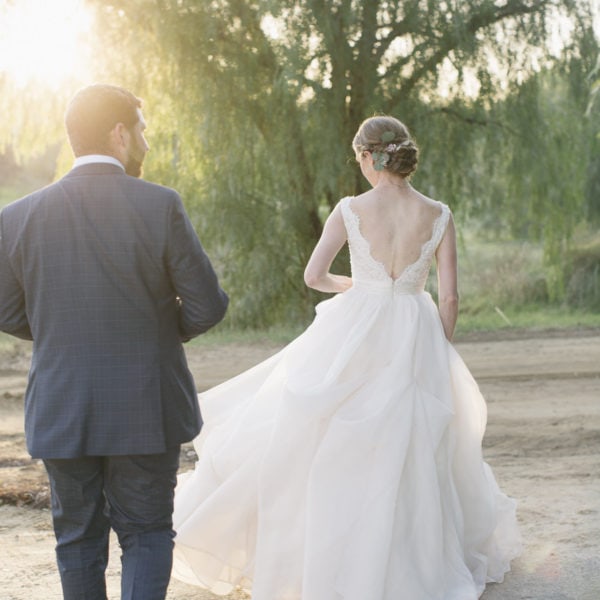 bride in a long white dress and groom in a blue suit, walking into a field at sunset